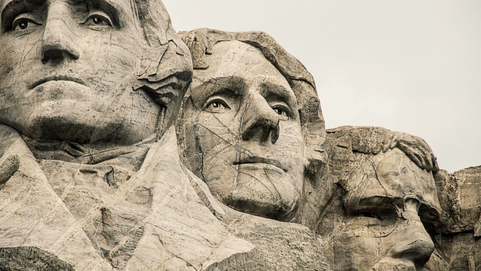 Close view of the carved stone faces of four U.S. presidents on Mount Rushmore with weathered rock details visible against a pale sky.