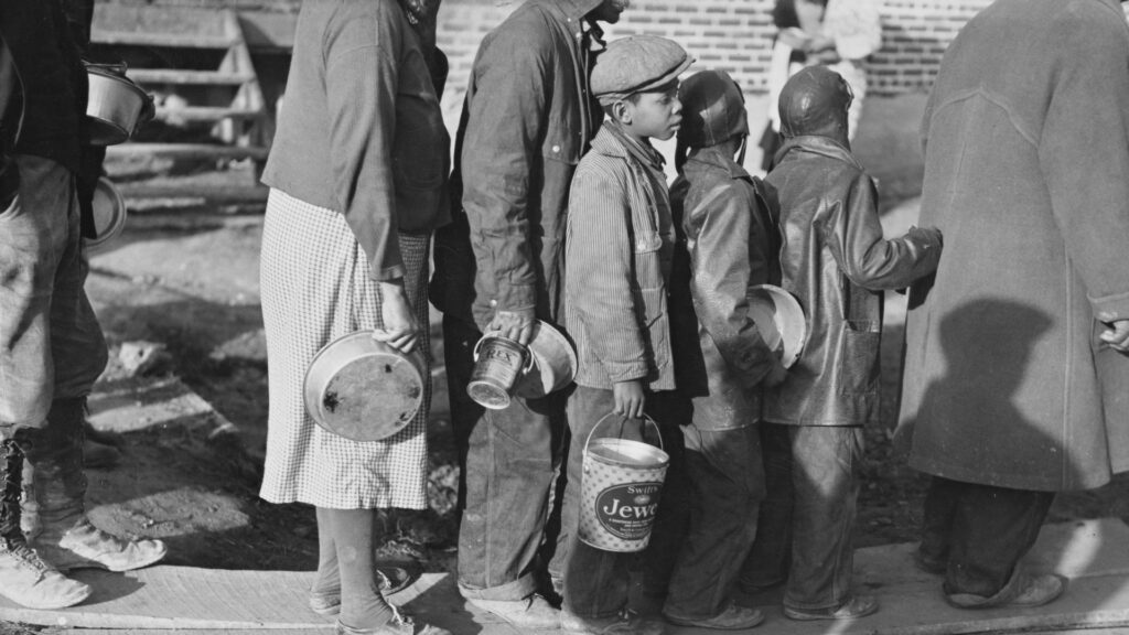 Black and white historical photograph of adults and children standing in line carrying empty pots buckets and containers. Several children wear caps and heavy coats while waiting with the group.