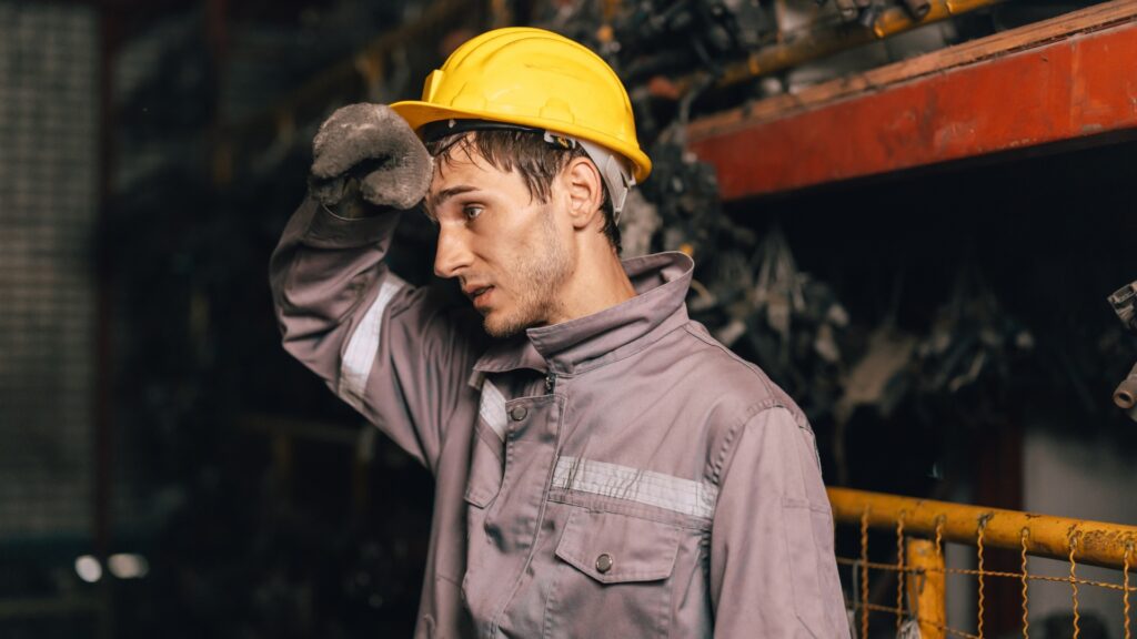 Factory worker wearing gray protective clothing and a yellow hard hat wipes his forehead with a gloved hand.