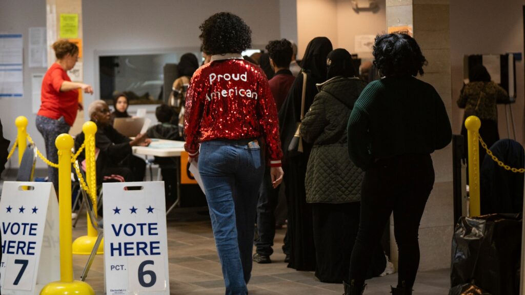 Voters wait in line inside a polling place beside signs reading "VOTE HERE PCT. 6" and "VOTE HERE PCT. 7." A person in a red sequined jacket reading "Proud American" walks toward the voting area.