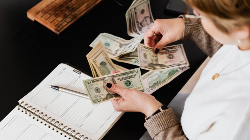 A woman counts U.S. dollar bills beside an open planner and pen on a desk.