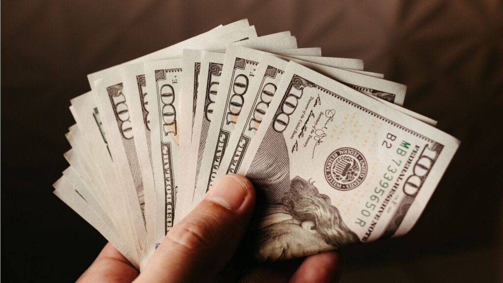 Close-up shot of a hand holding a fan of U.S. $100 bills against a dark background.