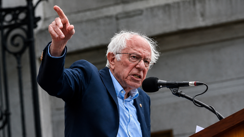Bernie Sanders speaking at a podium and pointing upward during a public address.