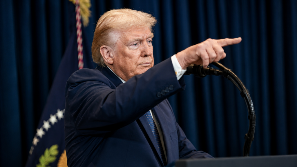 Donald Trump pointing while speaking at a podium with a U.S. flag behind him.