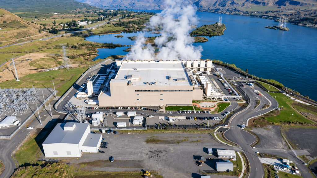 Large industrial facility beside a lake with steam rising from rooftop cooling equipment.