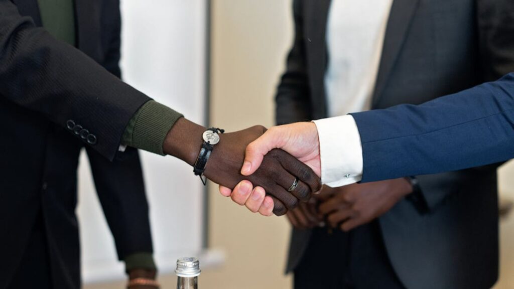 Two people in business suits shaking hands over a conference table, with another person standing in the background.