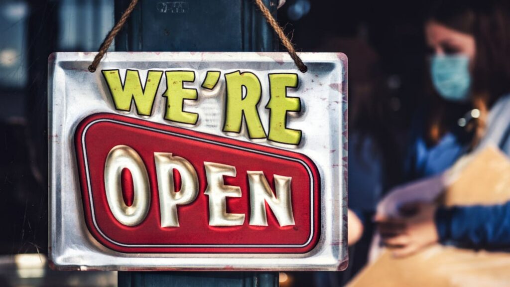 A 'We're Open' sign hanging on a shop door, with a masked person inside in the blurred background.