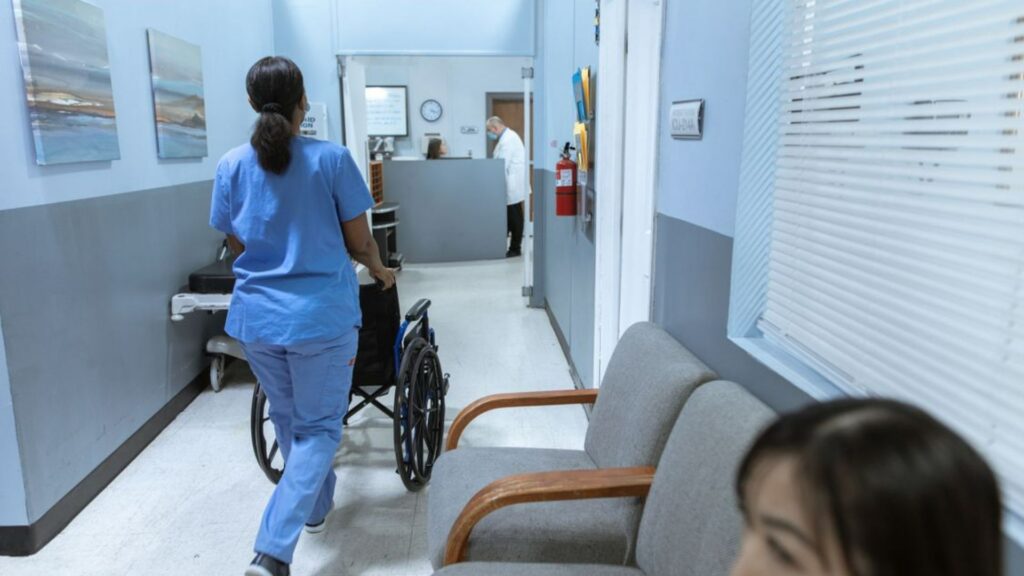 A nurse in scrubs pushes a wheelchair through a well-lit hospital corridor, attending to patient care.