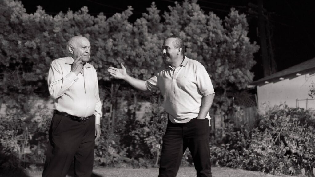 A father and son, sharing a candid moment in a garden setting, black and white photo.