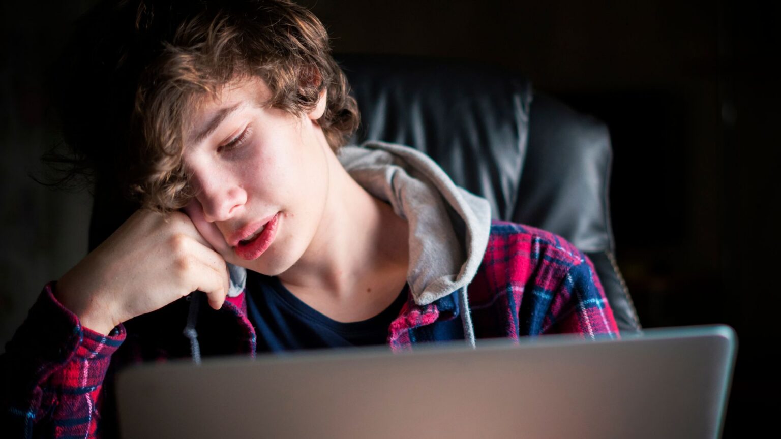 Young man with curly hair resting his head on his hand while looking tiredly at a laptop screen in a dimly lit room.