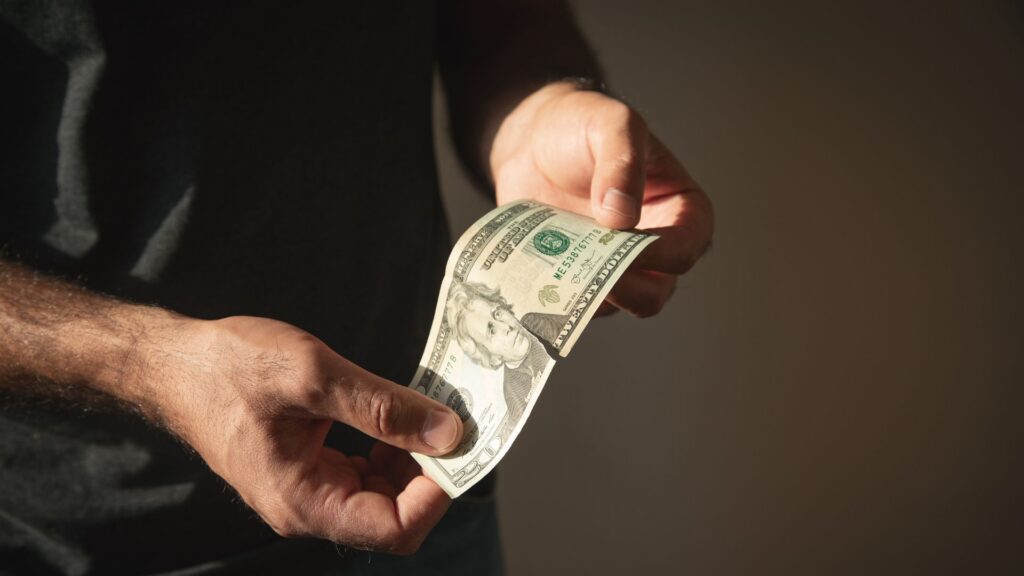 Person holding a slightly torn twenty dollar bill against dark background.