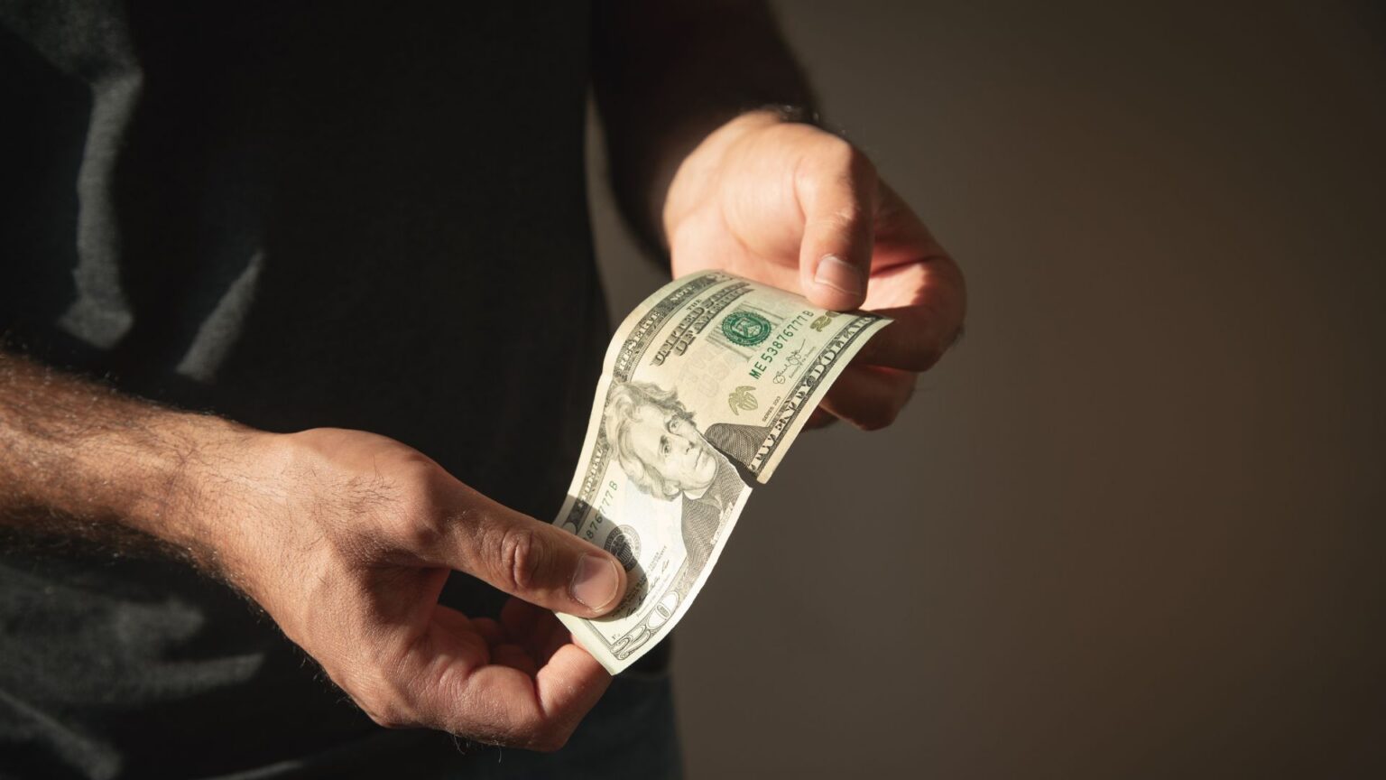 Person holding a slightly torn twenty dollar bill against dark background.