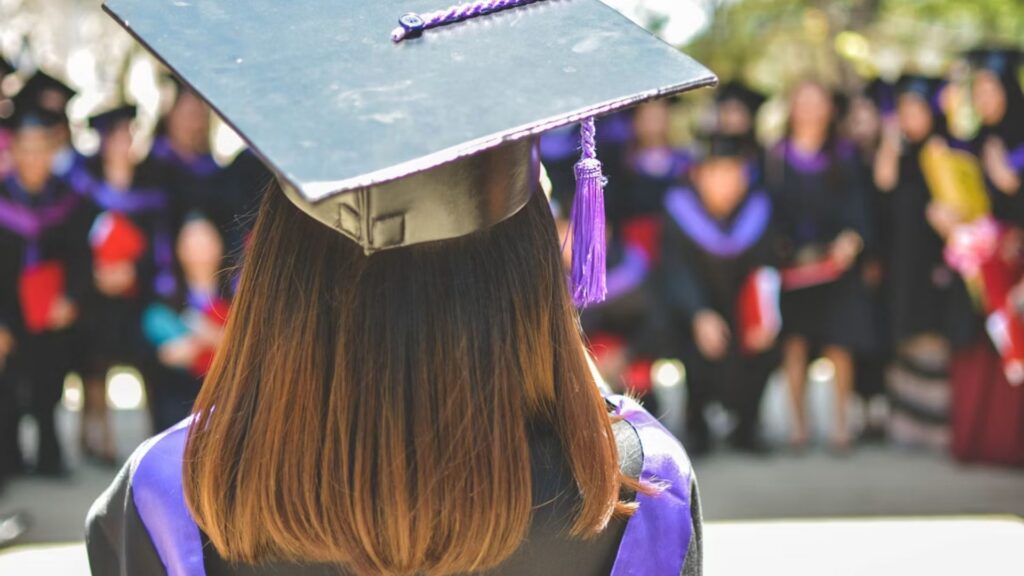 Rear view of a graduate wearing a black cap and purple-trimmed gown, facing a crowd of fellow graduates at an outdoor ceremony.