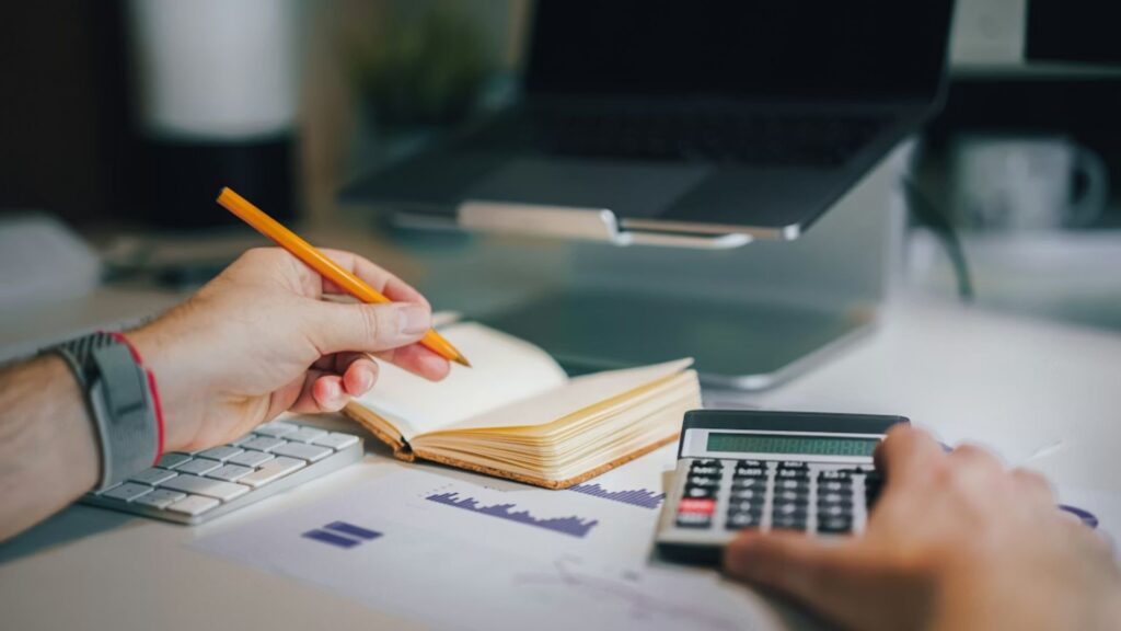 A person holding a pencil and calculator, reviewing financial documents at a desk.